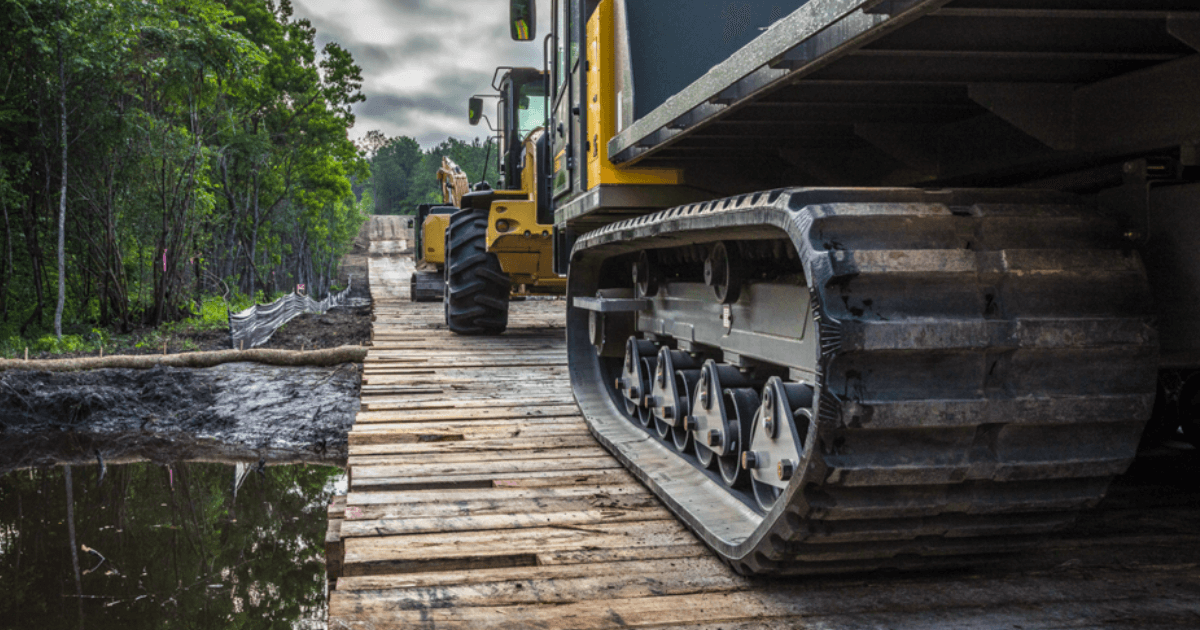 Construction vehicles driving on wooden Yak Access Mats across muddy water.